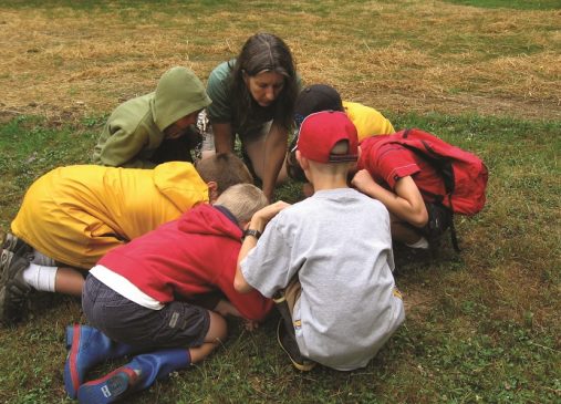 A group of children kneel around an instructor who shows them something in the grass at Shaker Lakes Nature Center