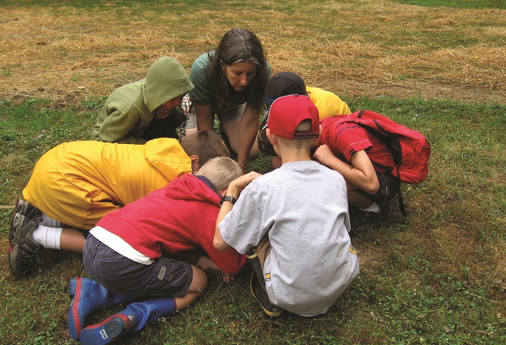 A group of children kneel around an instructor who shows them something in the grass at Shaker Lakes Nature Center