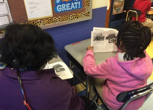 A woman sits with a young student as they read a book together