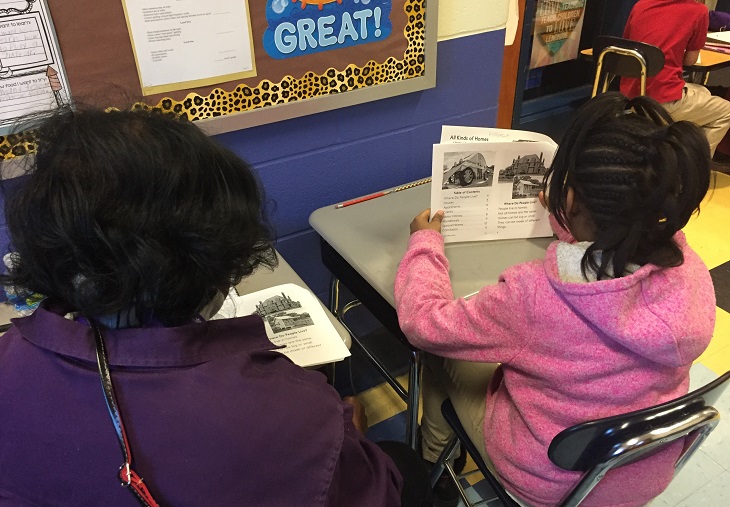 A woman sits with a young student as they read a book together