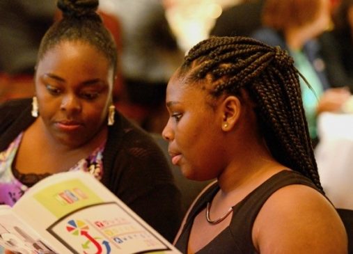 Two girls sit next to eachother reading a MyCom event program