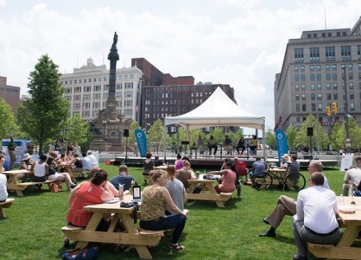 a crowd sits at picnic tables on public square listening to city club panel