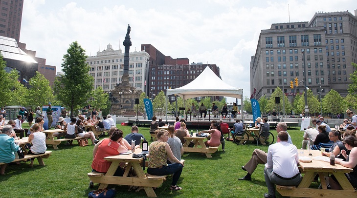 a crowd sits at picnic tables on public square listening to city club panel