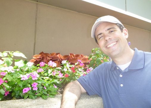 Frank Jason Feola stands smiling in front of flower bed