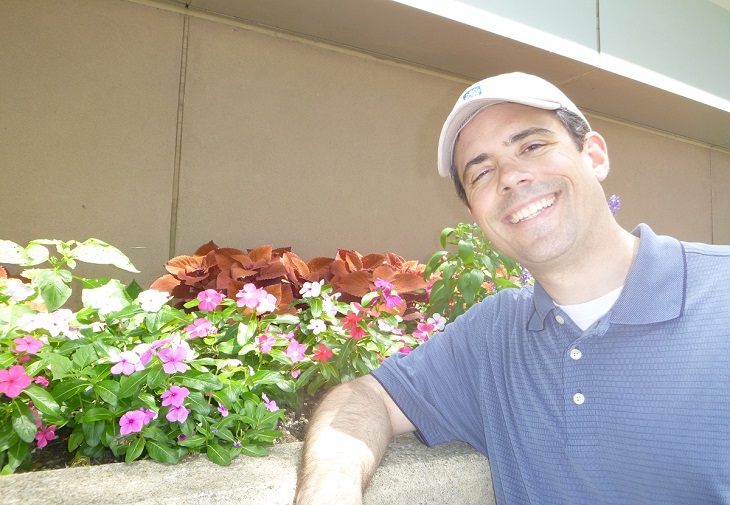 Frank Jason Feola stands smiling in front of flower bed