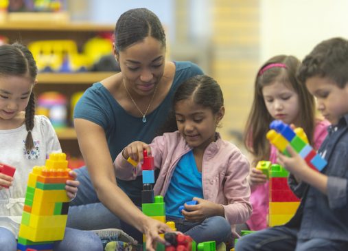 A teacher and her kindergarten students are playing with toy building blocks in their classroom.