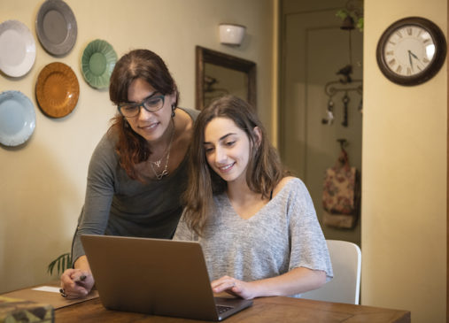 Woman leans over teenager seated at table on laptop