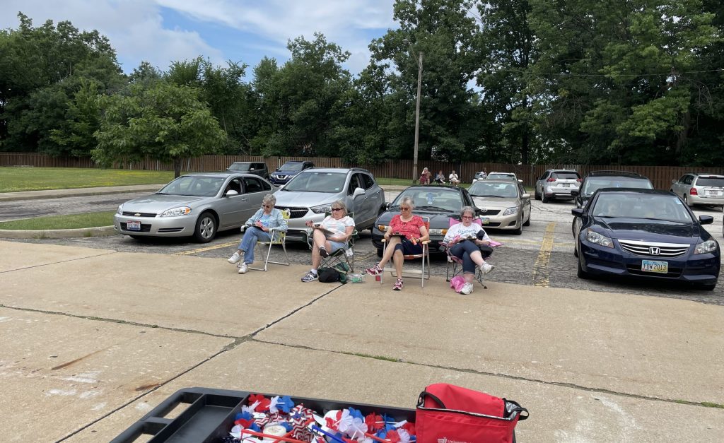 A group of women sit in folding chairs outside in a parking lot.
