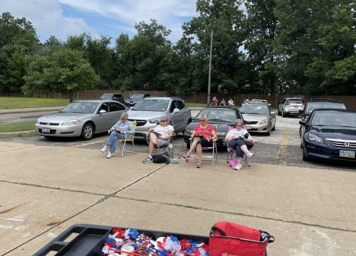 A group of women sit in folding chairs outside in a parking lot.