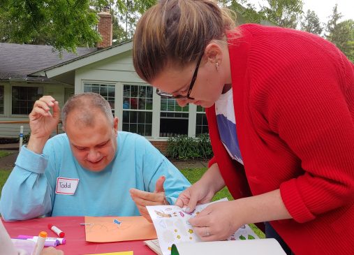 One person sits at a table while another leans over as they work on an activity together