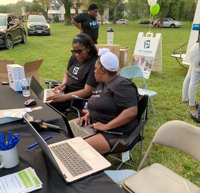 Two people sit behind a Pcs for People information table at a community event