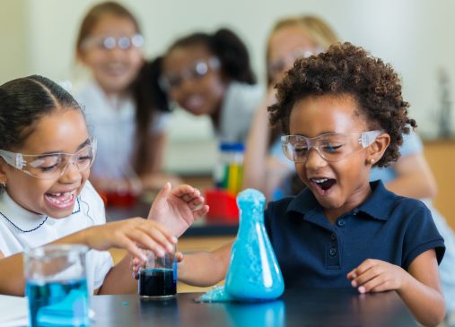 A group of young girls in a science lab sit in front of a beaker doing an experiment