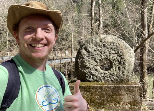 Ale stands outside giving a thumbs up in front of a stone wheel