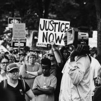 A black and white photo shows a group of protestors with one holding a "justice now" sign