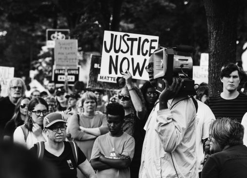 A black and white photo shows a group of protestors with one holding a "justice now" sign