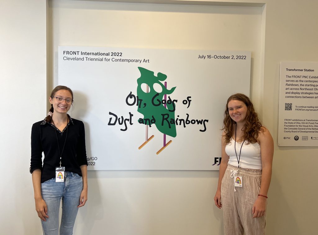 Audrey Ray and another intern stand in front of a wall sign for "Oh Gods of Dust and Rainbows" 2022 FRONT Exhibition